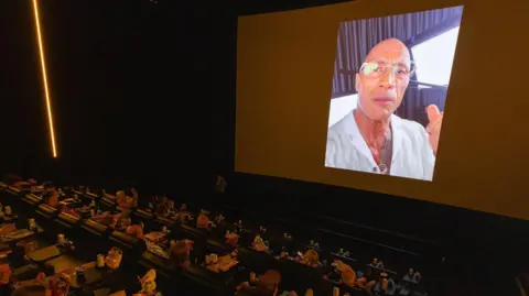 James Gillham/Disney A selfie-style video of The Rock on a cinema screen as a packed theatre of children watch. 