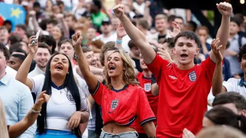 England fans celebrate in a pub garden. A crowd of men and women, in the foreground are two women and two men with their arms up wearing football t-shirts.