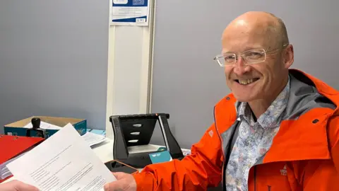 The Kendal Green Party Phil Clayton handing in a his nomination papers for the 2024 General Election. He is sitting at an office desk handing over the papers, which are being taken by another person. He is bald and wears rimless glasses and an orange waterproof coat. He is smiling widely at the camera.