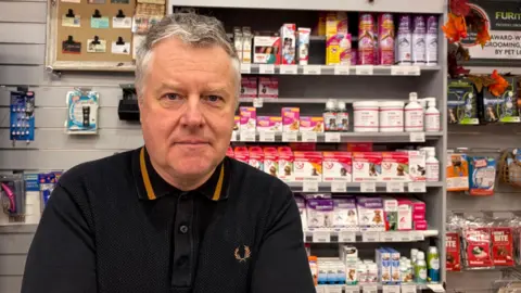 A man with short grey hair is looking into the camera. He is wearing a dark polo shirt. Behind him is a shop display.