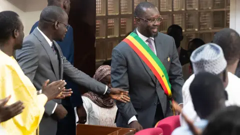 AFP via Getty Images Ousmane Sonko wears a sash in parliament. He smiles as supporters applaud him.