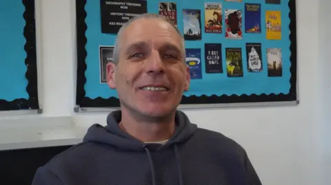 A man with short greay hair smiles at the camera. He is wearing a dark-coloured hooded top. In the background is a bright blue display about books on a school wall.