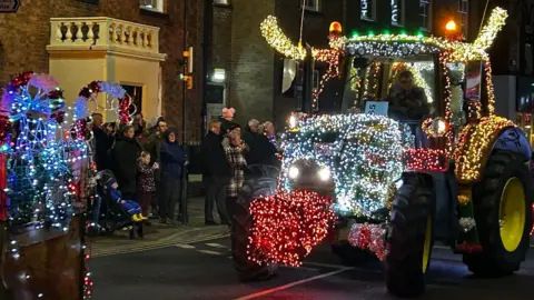 A tractor is brightly-lit with reindeer antlers and a red nose and driving through Oswestry. People are standing on the pavements watching and the tail end of another vehicle with fairy lights can be seen. The tractors are passing a building with a decorative entrance.