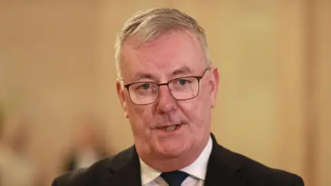 PA Media Mike Nesbitt speaking in Stormont Hall. He is wearing a black suit, white shirt and blue tie. He has dark framed glasses on. 