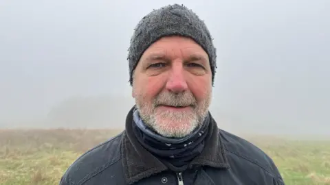 Carl Pickup looking down the camera, he's wearing a navy jacket and a grey woollen hat. Behind him you can see some of the grassland through the fog, with a copse of trees just visible over his right shoulder. 