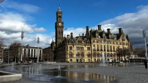 A Victorian Gothic-style building has a clock tower and large arched windows. Outside are a public square and fountains.