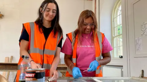 Rebecca Austin and Karishma Nanhu wearing safety glasses and high-visibility vests repair and re-putty a historic window frame during a heritage conservation project at Wentworth Woodhouse.