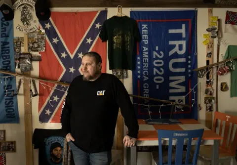 Joel Gunter/BBC File image of Michael Macartney at his home in Virginia. He stands in front of a wall decked out with various flags
