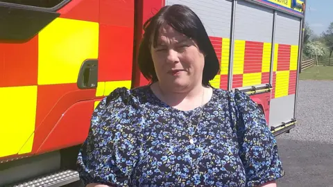 A woman is standing in front of a fire engine. One hand is folded over the other. Fields are in the background.