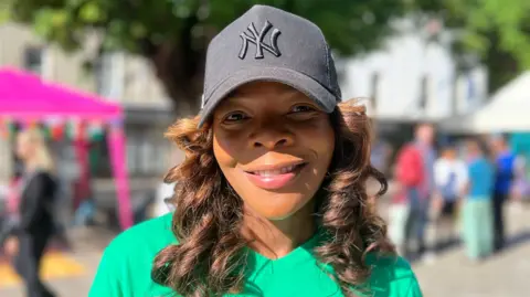 Close-up portrait of a Lainah who is wearing a black baseball cap with a embroidered “NY” logo and a green T-shirt, standing outdoors in a sunlit public space with trees, tents, and blurred people in the background