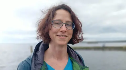 Nicole smiling at the camera. She is wearing a blue top with a green jacket over it. She is wearing green/blue glasses and has short brown hair.