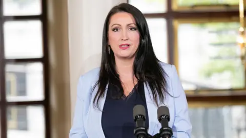 Emma Little-Pengelly wearing a light blue blazer and navy top, she has black hair
