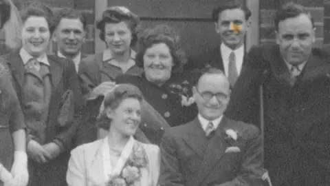 Pauline Tasker A black and white image of Peggy and Len Tasker on their wedding day. She is wearing flowrs on her dress and he has a dark suit on. They are surrounded by wedding guests 
