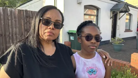 BBC Angela with her 11-year-old daughter Ava. They are both wearing sunglasses on a pleasant day. Angela has long black hair, black sunglasses and a black top on. Ava has black hair tied up, black sunglasses and is wearing a pink vest top with a Disney character on.