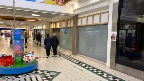 Pacemaker Inside of Connswater Shopping Centre, showing floor tiles and windows - empty units are seen with shoppers walking past them