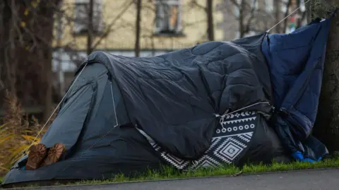 Getty Images Dark blue tent on grass with a a pair of shoes sticking out of the entrance with only the soles showing. There are trees and buildings in the background.