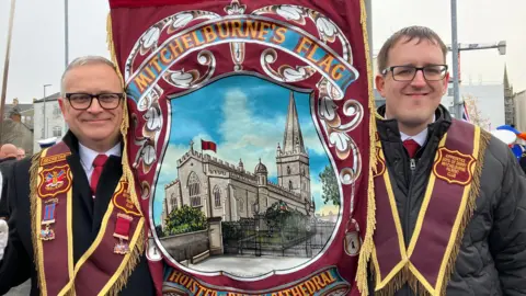 Two men in collarettes stand either side of a banner showing St Columbs Cathedral in Derry. They are taking part in an Apprentice Boys parade