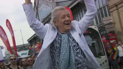 Screengrab of a video of Sir Ian McKellen at Manchester Pride in 2015. He stands in front of a crowd with his arms raised in celebration