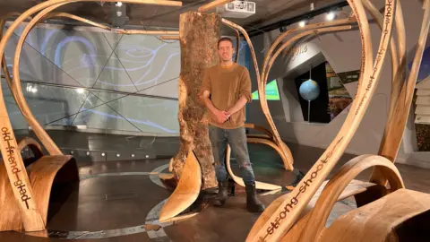 Artist Charlie Winney stands next to his sculpture. He is clean shaven with short dark hair and is wearing a brown jumper and blue jeans. The sculpture consists of a tree trunk with pieces of bent wood coming form it connecting to benches