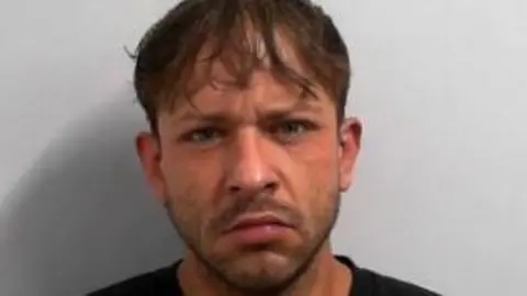 A mugshot of a man frowning directly at the camera in a police mugshot. He has a short dark beard and medium length brown hair. 