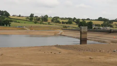 Nick Coggins A reservoir with low water levels, large stretches of the reservoir basin are dry around the water. A tower is in the middle and its base is submerged. Construction machinery is working on the opposite side of the reservoir.
