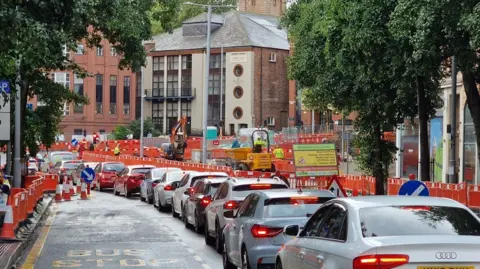 Local Democracy Reporting Service A queue of at least 10 cars displaying red brake lights wait on Upper Parliament Street in Nottingham on the approach to the new T-junction on Maid Marian Way. Construction barriers, traffic cones and building machinery can be seen in the background.