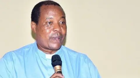 Fr Charles Kitima in a blue clerical shirt and white collar and holding a microphone