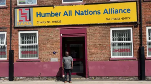 Kevin Shoesmith/BBC Francis Ahiakpa stands outside the Humber All Nations Alliance, a two-story brick building with purple facades and a large yellow sign with the organization's name printed in bold blue lettering. 