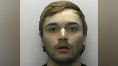 A police mugshot of Jake Ahmed. He has wavy brown hair and a vacant expression with his mouth slightly agape.
