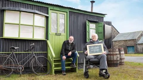 Alistair Corbett Seimon and his dad Stephen sat outside their mobile museum. Seimon is sat on the steps, while Stephen sits in his wheelchair to the right. He is holding a sign saying Iscoed Bakery. This side of the museum is a replica of Seimon's great-grandmother's bakery.