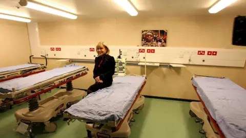 PA Media Dame Ann Gloag sitting on a bed on the Africa Mercy hospital ship, with several other beds nearby.