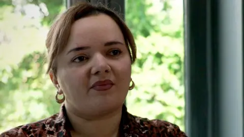 Issy Vine has brown hair and is wearing a brown shirt with a leopard print pattern. She is sitting in a kitchen with some green trees outside the window behind her.