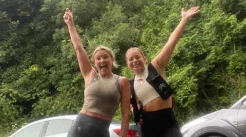 Andrea McCartney Andrea McCartney with her friend Ruth Busby in their running gear as they prepare for Andrea's marathon challenge. They are standing side by side and each have one hand in the air, looking happy.