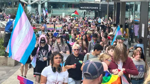 A crowd of people snakes down a city centre precinct, waving rainbow flags. Shops including Waterstones can be seen behind.