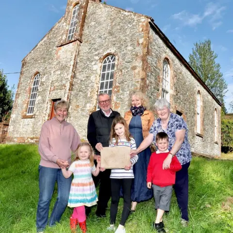 Bryan Armstrong A group of four older people and three children stands in front of Wamphray Church on a sunny day