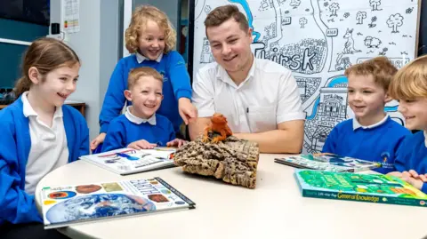North Yorkshire Council Ben McAuley and a number of primary-age pupils sit around a white table, with a model of a lizard on a log in the middle of the table. Mr McAuley wears a short sleeved white shirt. He has short brown hair. The pupils wear blue sweatshirts and white polo shirts. A number of science books are also on the table.
