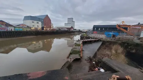 Andrew White The photo show the rather industrial setting of the River Hull, with concrete steps and discarded rubbish. An old rusty and broken foot bridge is covered by moss. The water is dirty and brown.
