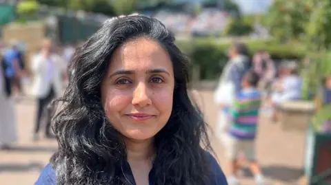 Anjum Gul is standing in the sunshins with spectators milling around behind her. She is wearing a blue top and has long black hair.