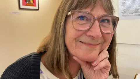 Trix Jones, who has shoulder-length hair and glasses, looking thoughtfully at the camera, smiling.