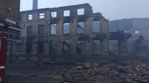 A burned down mill with rubble in the street and the outside frame remaning