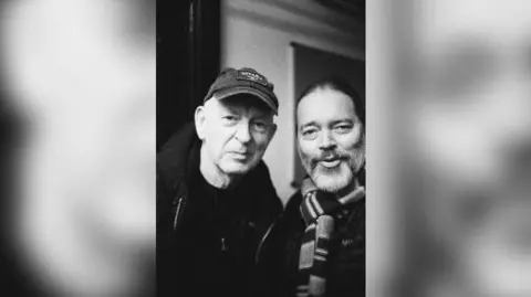 Conal Dodds A black and white portrait image of Conal Dodds (R) pictured with Alan McGee (L). Alan is wearing a baseball cap and Conal is wearing a striped scarf. They're both smiling at the camera.