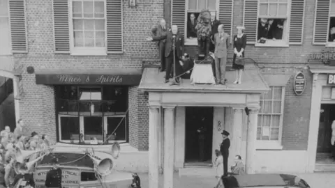 Getty Images A black and white image of the balcony of the former Red Lion Hotel, High Wycombe, Winston Churchill addresses a crowd of 8,000 citizens, at the start of his election campaign tour. There is a car to the left, people standing under the balcony, and people handing out windows at the top. 
