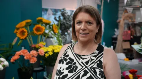 A woman with short brown hair is stood in front of flowers in her shop. She is wearing a black and white dress and looking straight into the camera.