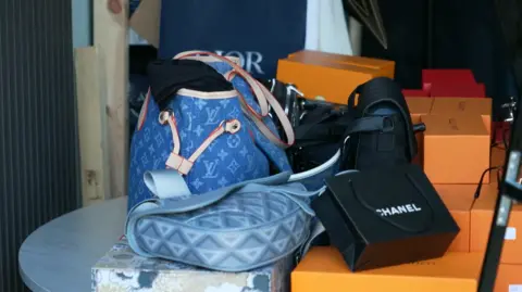 Imitation Louis Vuitton Neverfull handbag in denim next to many imitation LV orange sunglasses boxes and a black Chanel bag. 