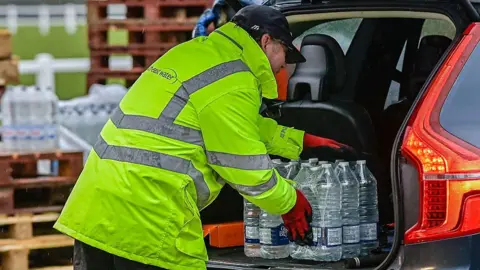 A man in a yellow hi-vis jacket putting in a pallet of water into the back of a red car.