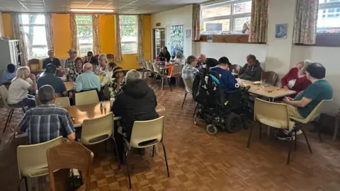 Cad Taylor, BBC A large room with a brown parquet floor. Lots of tables are set about with groups chatting in each