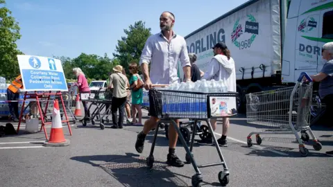 PA Media A man in a white shirt and shorts is pushing a trolley full of water bottles. People are queuing behind him and a lorry is in the background.