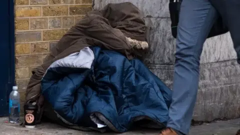 Getty Images Homeless person with coat on and sleeping bag while someone walks past