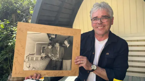A man with gray hair wearing glasses smiles as he holds an old black and white photograph on a cardboard mount with signatures around it. The photo shows him as a child with Graham Taylor, a dark-haired man wearing a blazer and tie, and a taller man with a dark mullet wearing a tie and jumper.