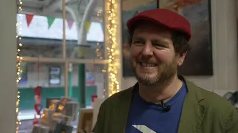 BBC/Emma Shales Paul stands in a green jacket and blue T-shirt with the national rail logo, as well as a red flat cap. He is standing in his gallery with artwork and fairy lights in the background.